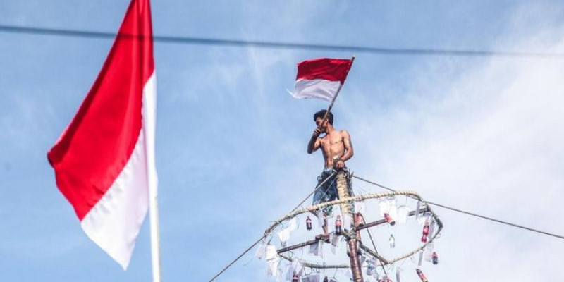 Seorang peraih juara lomba panjat pinang mengibarkan Bendera Merah Putih di puncak kemenangannya/ Net